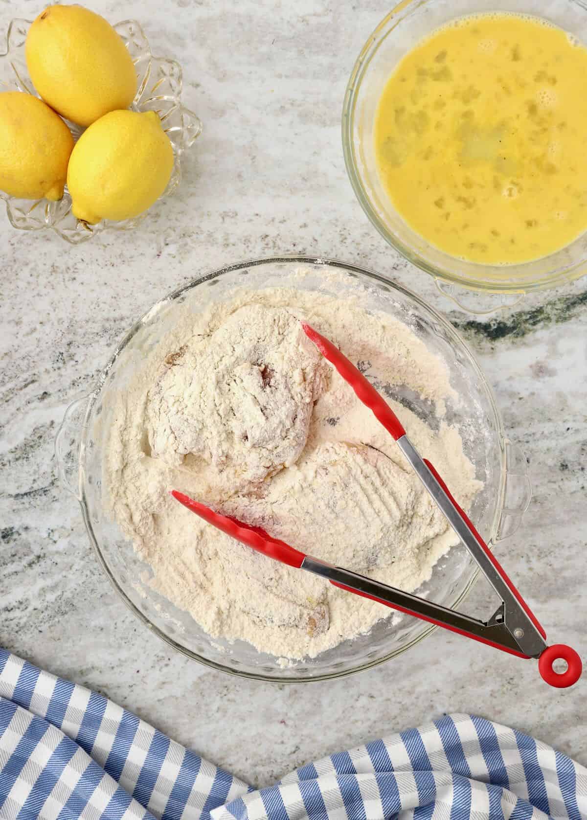 Dredging a chicken thigh in a bowl of flour using tongs. 