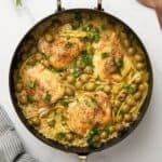 A large skillet with baked chicken and orzo in a sauce made with chicken stock and wine, with olives and strips of lemon.