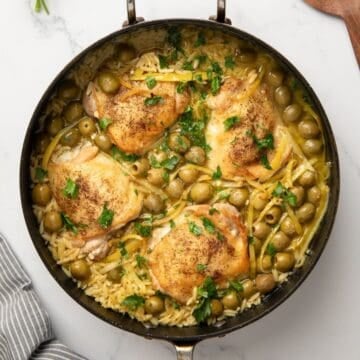 A large skillet with baked chicken and orzo in a sauce made with chicken stock and wine, with olives and strips of lemon.