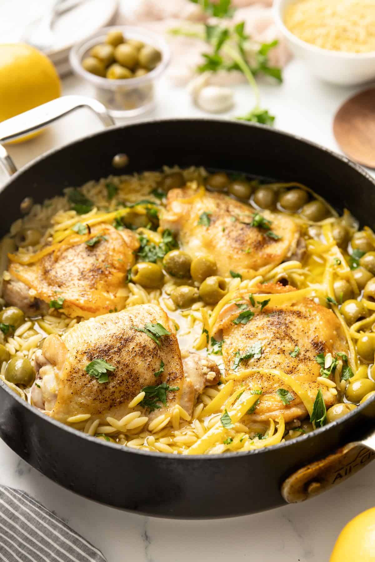 Pan-seared chicken in a skillet with cooked orzo, olives, lemon strips and parsley.  