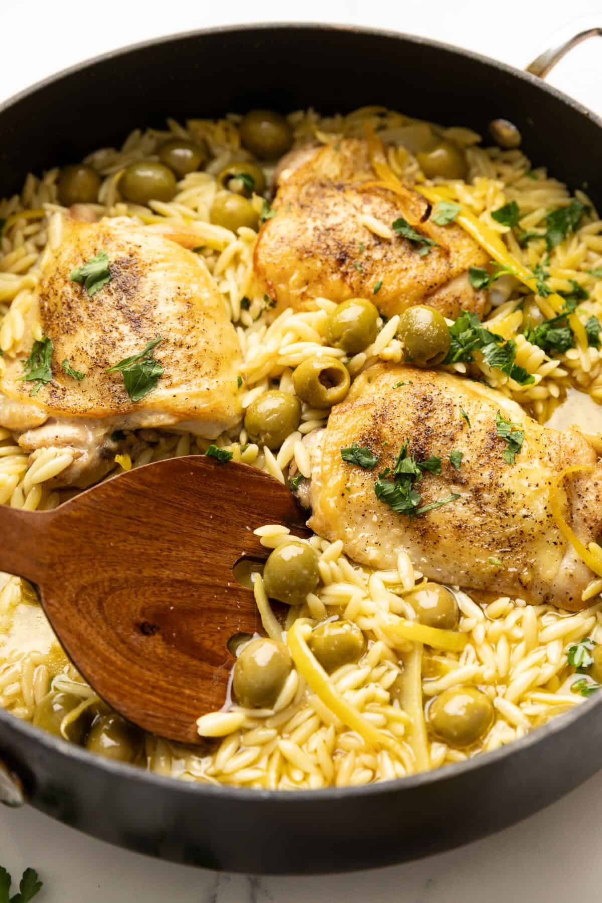 A skillet with chicken and orzo, olives, and lemons with a large wooden spoon.