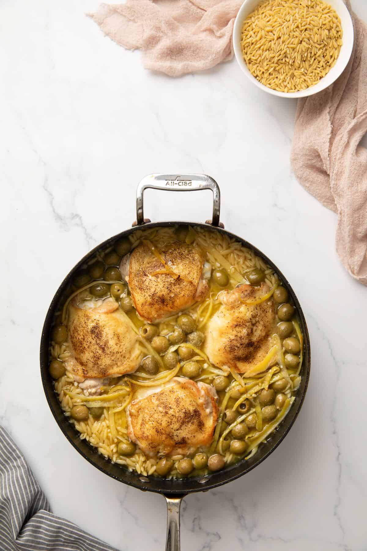 A large skillet full of pan seared chicken thighs, orzo, strips of lemon peel, and chicken stock. 