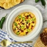 A white Crock-Pot full of Jalapeno Popper Dip topped with melted cheese, bacon, and sliced jalapenos.