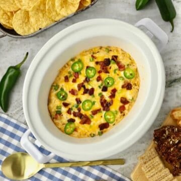 A white Crock-Pot full of Jalapeno Popper Dip topped with melted cheese, bacon, and sliced jalapenos.