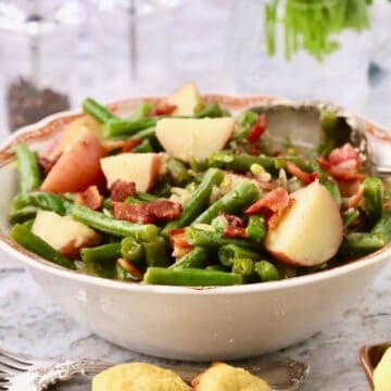 A white serving dish with cooked green beans and red potatoes garnished with crispy pieces of bacon.