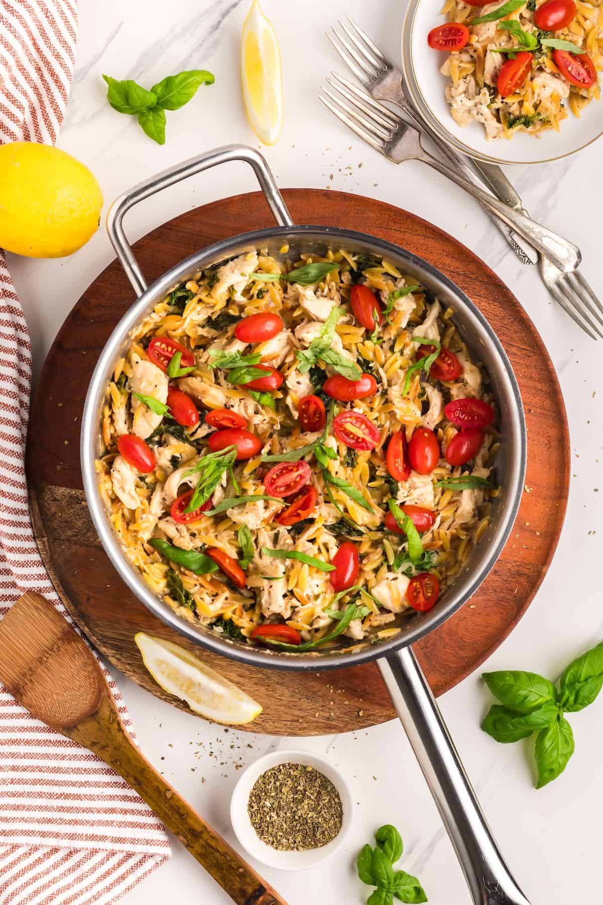 A skillet is full of lemon chicken orzo pasta, garnished with sliced cherry tomatoes and basil. 