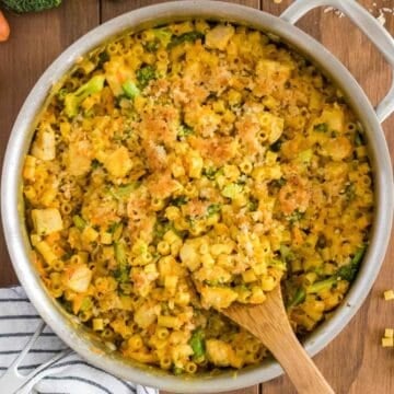 A skillet full of cheesy chicken broccoli pasta with a wooden spoon.