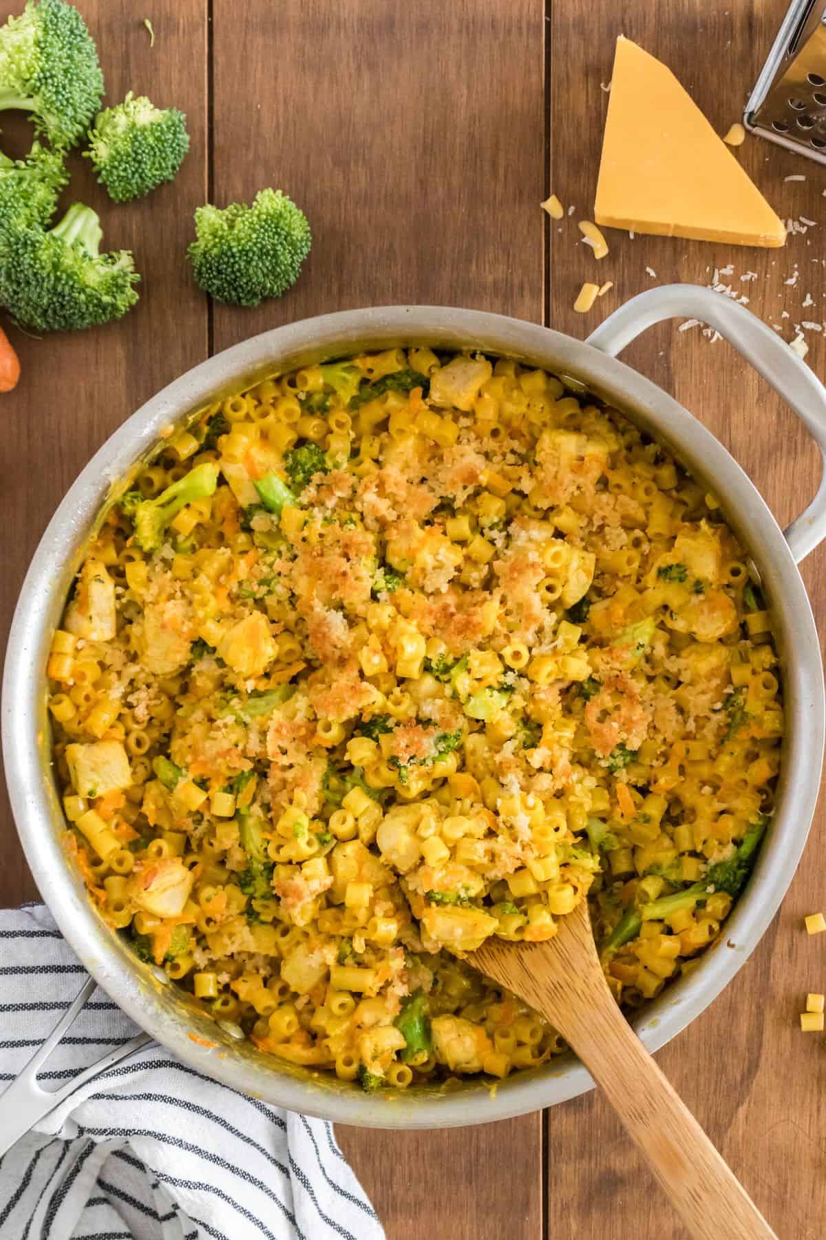 A skillet full of cheesy chicken and broccoli pasta with a wooden spoon.