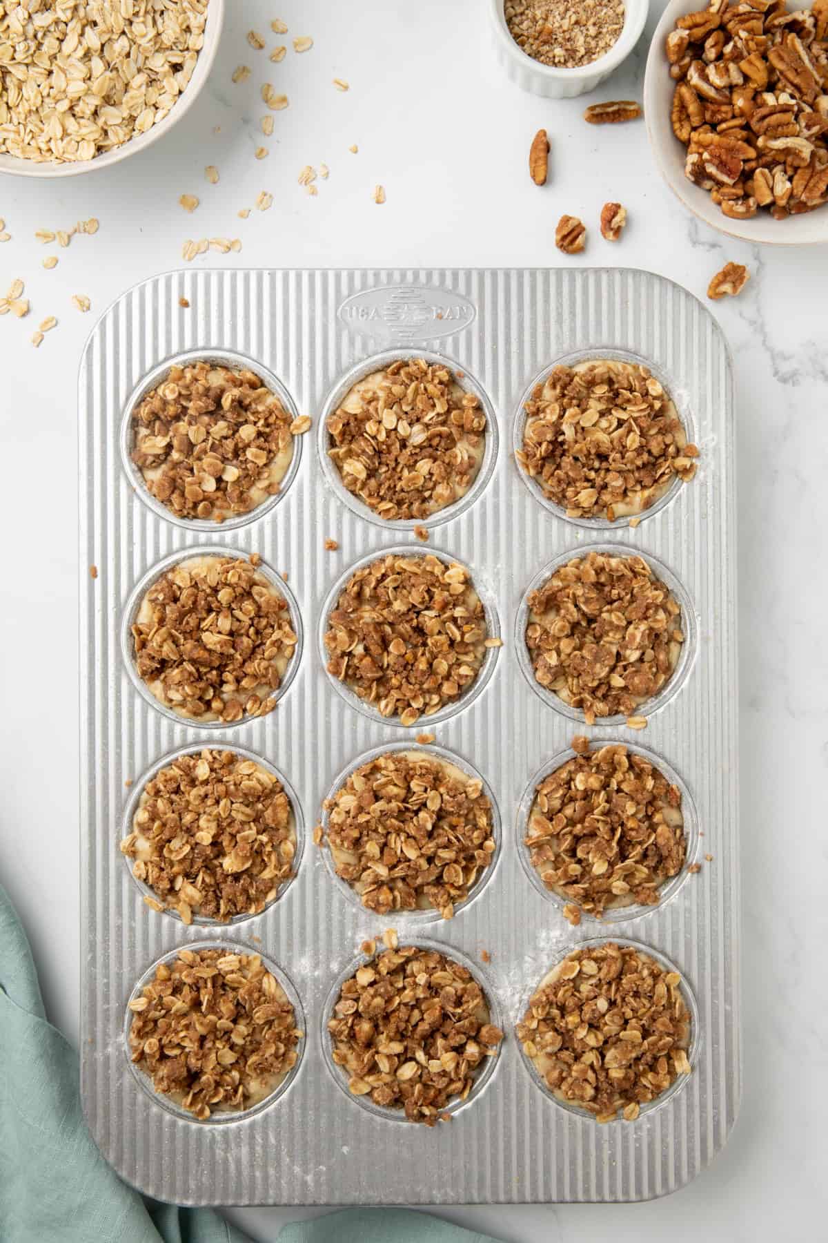 A muffin pan with oatmeal muffin batter topped with an oat and pecan crumble. 