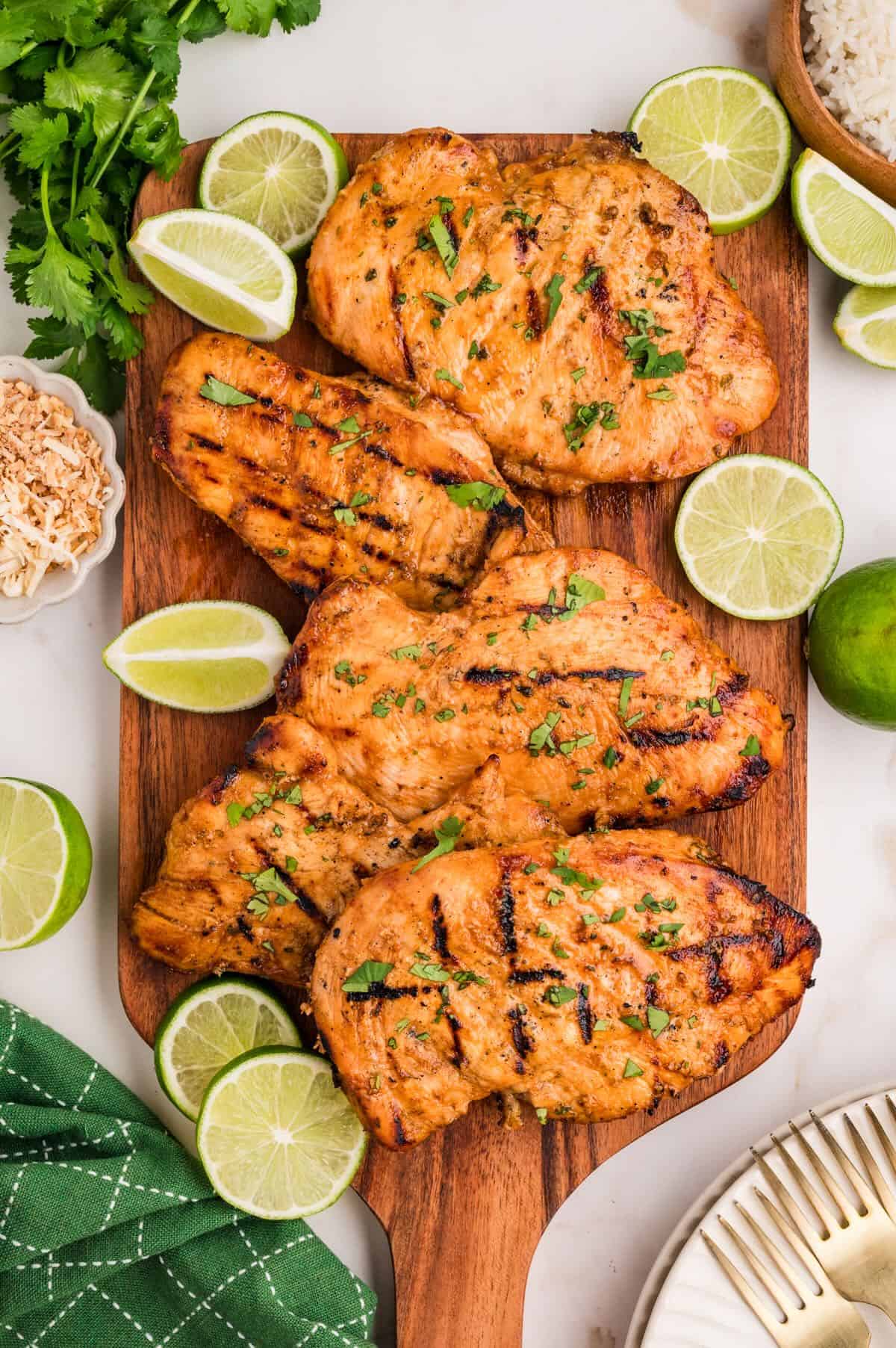 A cutting board piled high with grilled coconut lime chicken breasts, garnished with limes. 