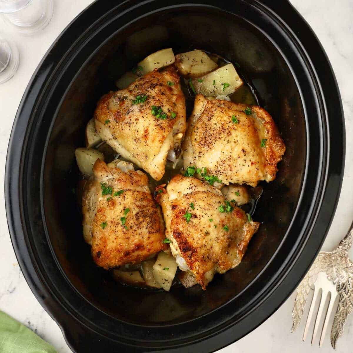 Cooked potatoes and chicken thighs in a black crock pot bowl.