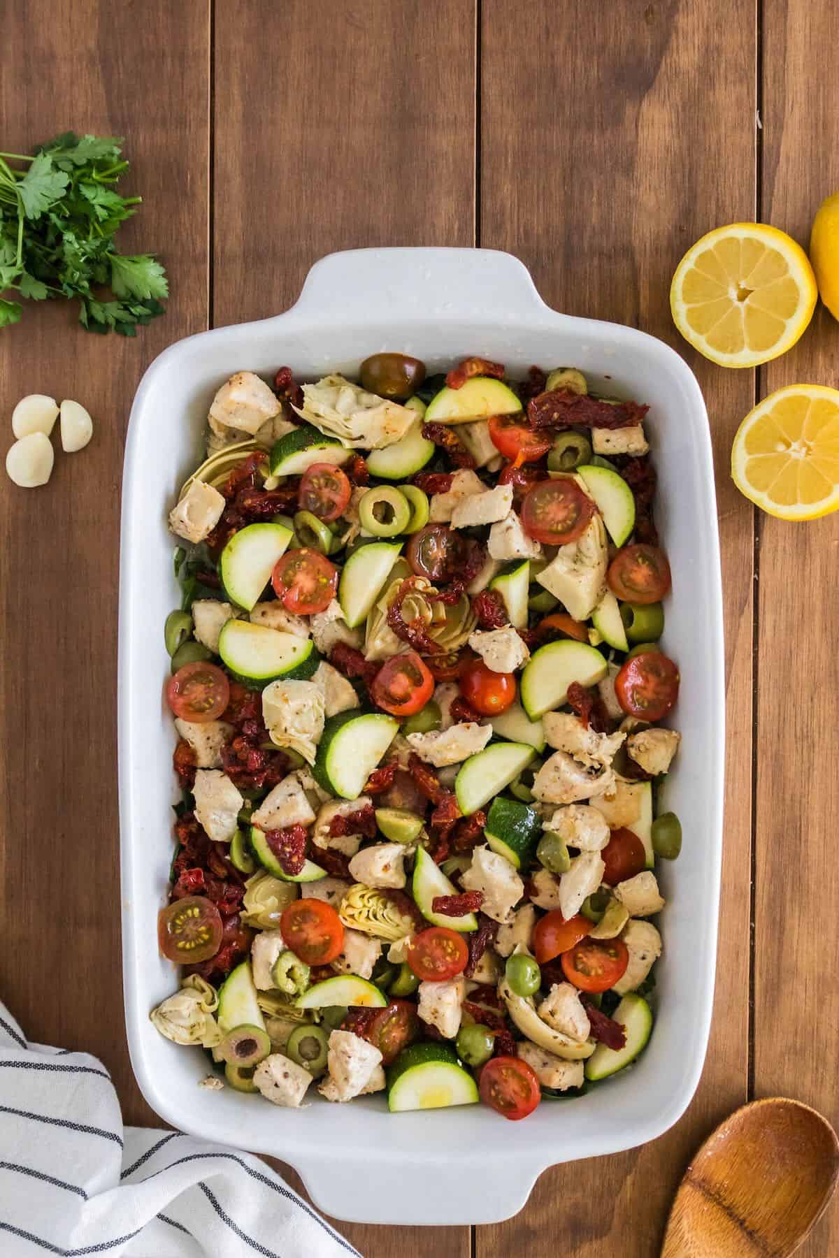 Chicken, tomatoes, zucchini, olives, sun-dried tomatoes, and artichokes in a baking dish. 