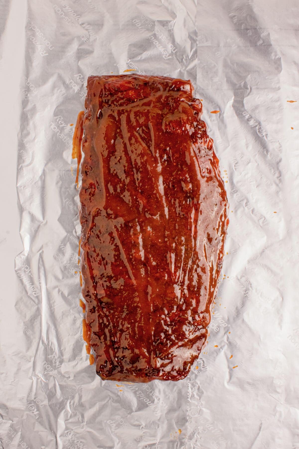 A rack of cooked spare ribs topped with BBQ sauce on a sheet of foil, 
