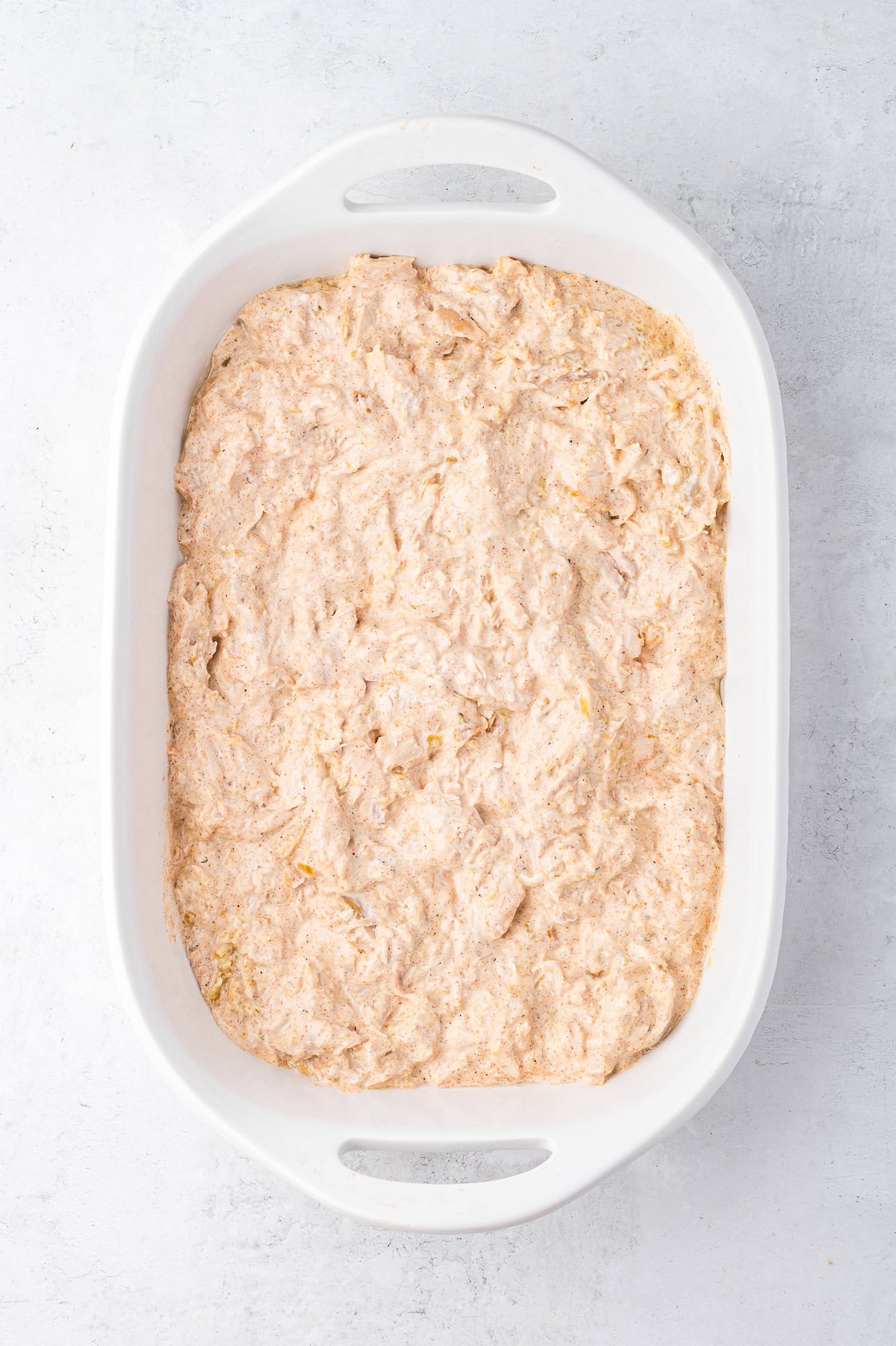A white ceramic baking dish with a chicken, salsa verde, and sour cream mixture spread out on the bottom.
