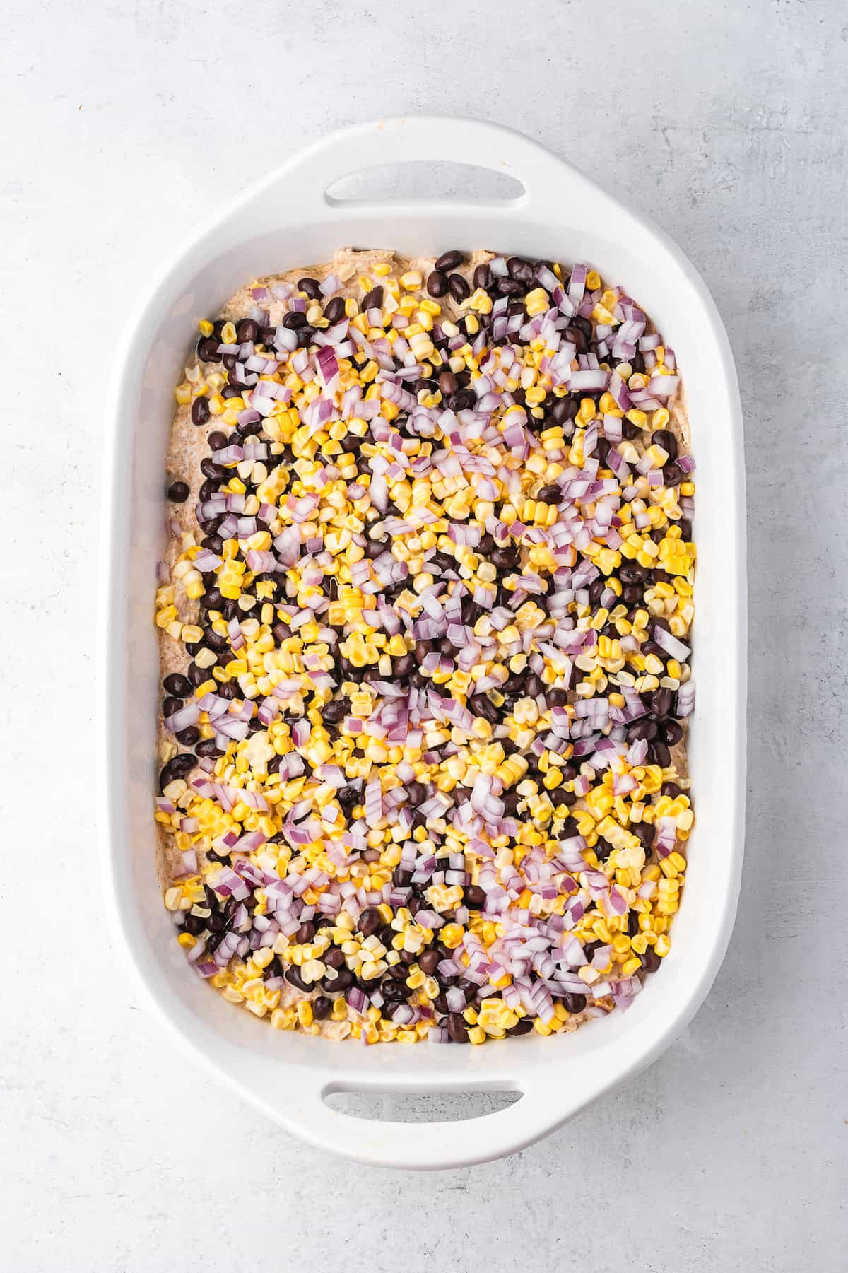 A white ceramic baking dish with layers of chicken mixture, black beans, corn, and red onion. 