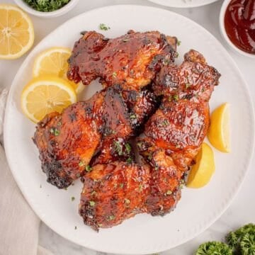 A white plate full of grilled BBQ chicken thighs with BBQ sauce, garnished with sliced lemons.