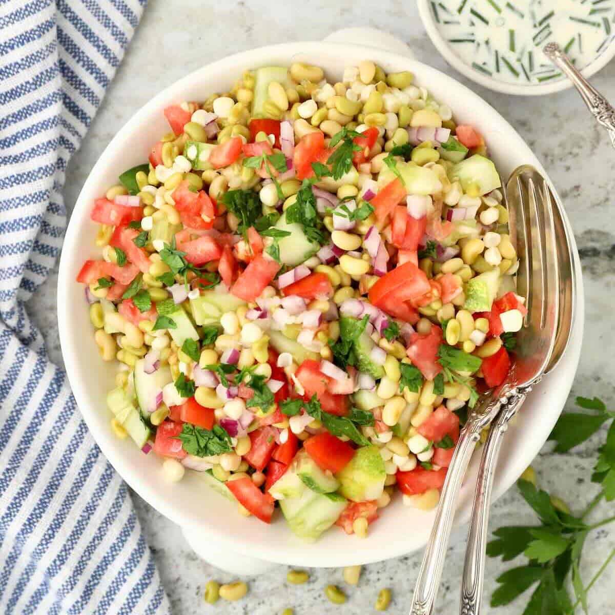 A white bowl full of Southern Pea Salad made with white acre peas, chopped tomatoes, cucumber, and corn, plus a bowl of buttermilk dressing.