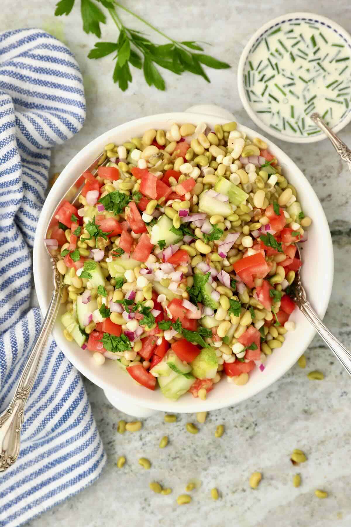 A bowl full of simple pea salad with white acre peas with two serving forks and a blue and white striped napkin. 