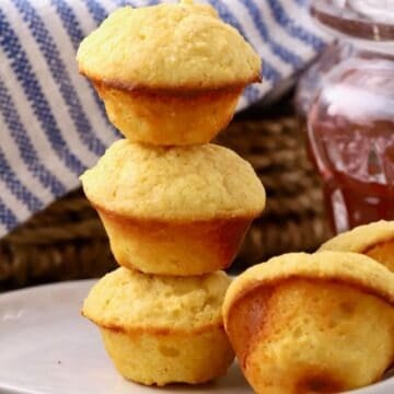 Three mini cornbread muffins stacked on a plate with two other muffins, with a jar of jelly next to it.