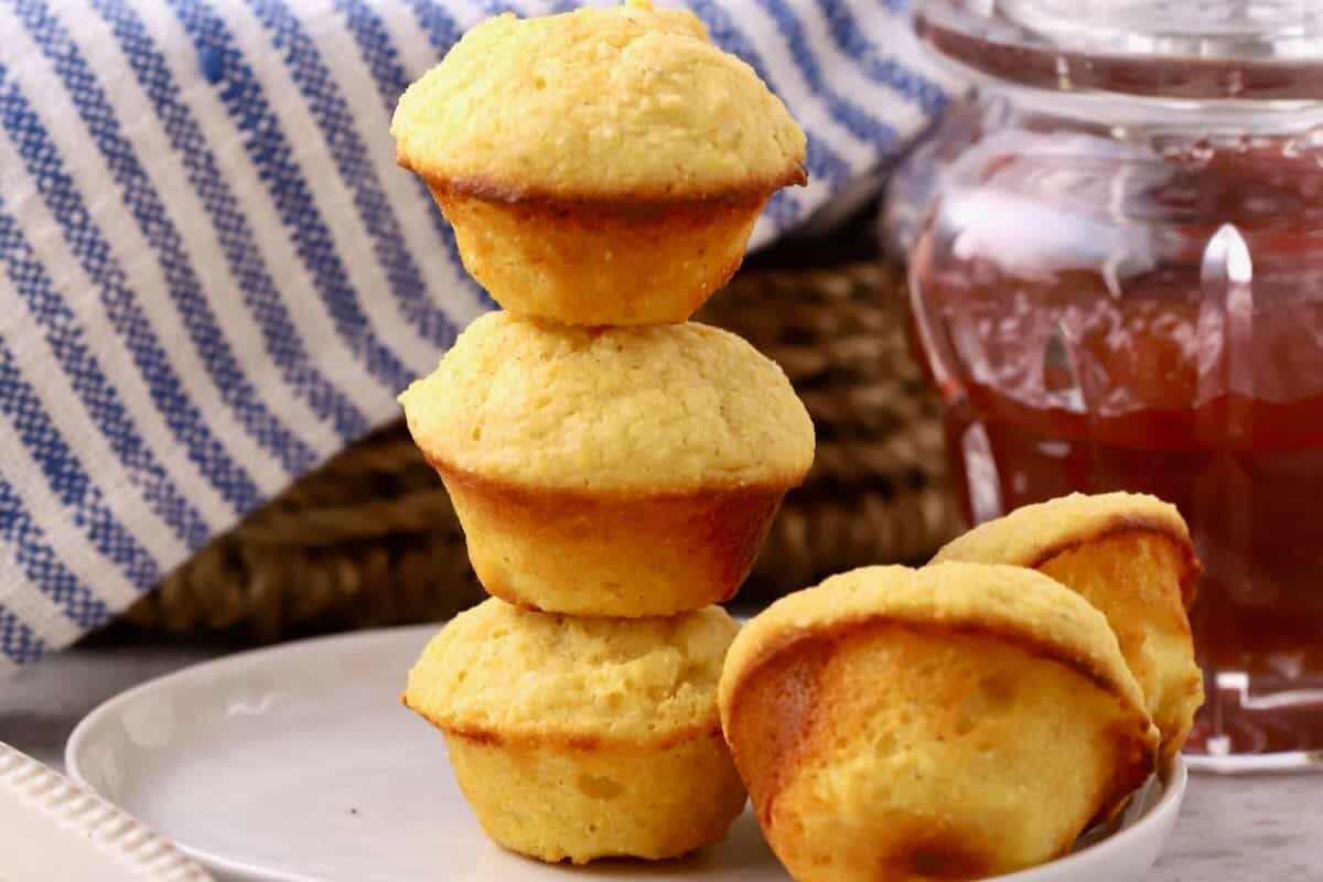 Three mini cornbread muffins stacked on a plate with two other muffins, with a jar of jelly next to it. 