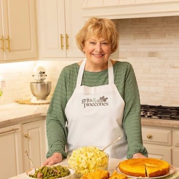 Sharon Rigsby with a white apron with the grits and pinecones logo in front of a platter of fried chicken potato salad and cornbread. 