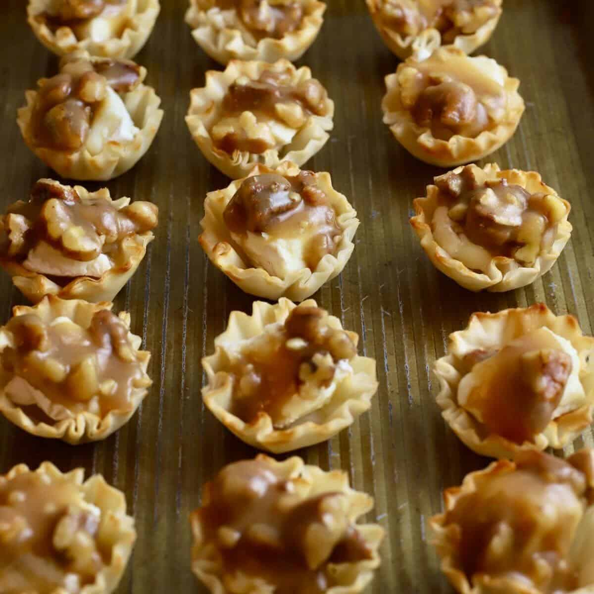 A baking sheet with phyllo cups with cubes of brie cheese, topped with a pecan, bourbon, and butter mixture. 