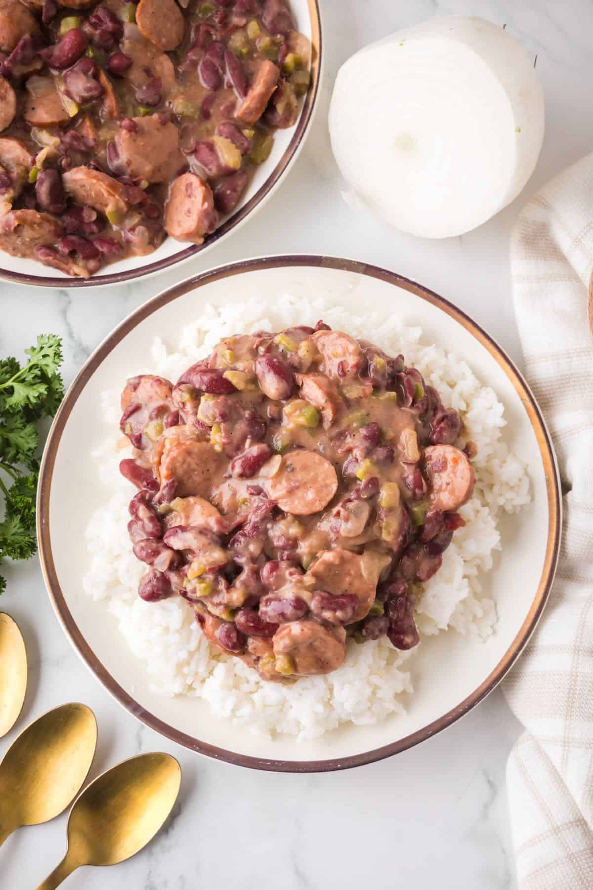 A white plate with a gold rim with white rice topped with red beans with sausage and the holy trinity. 