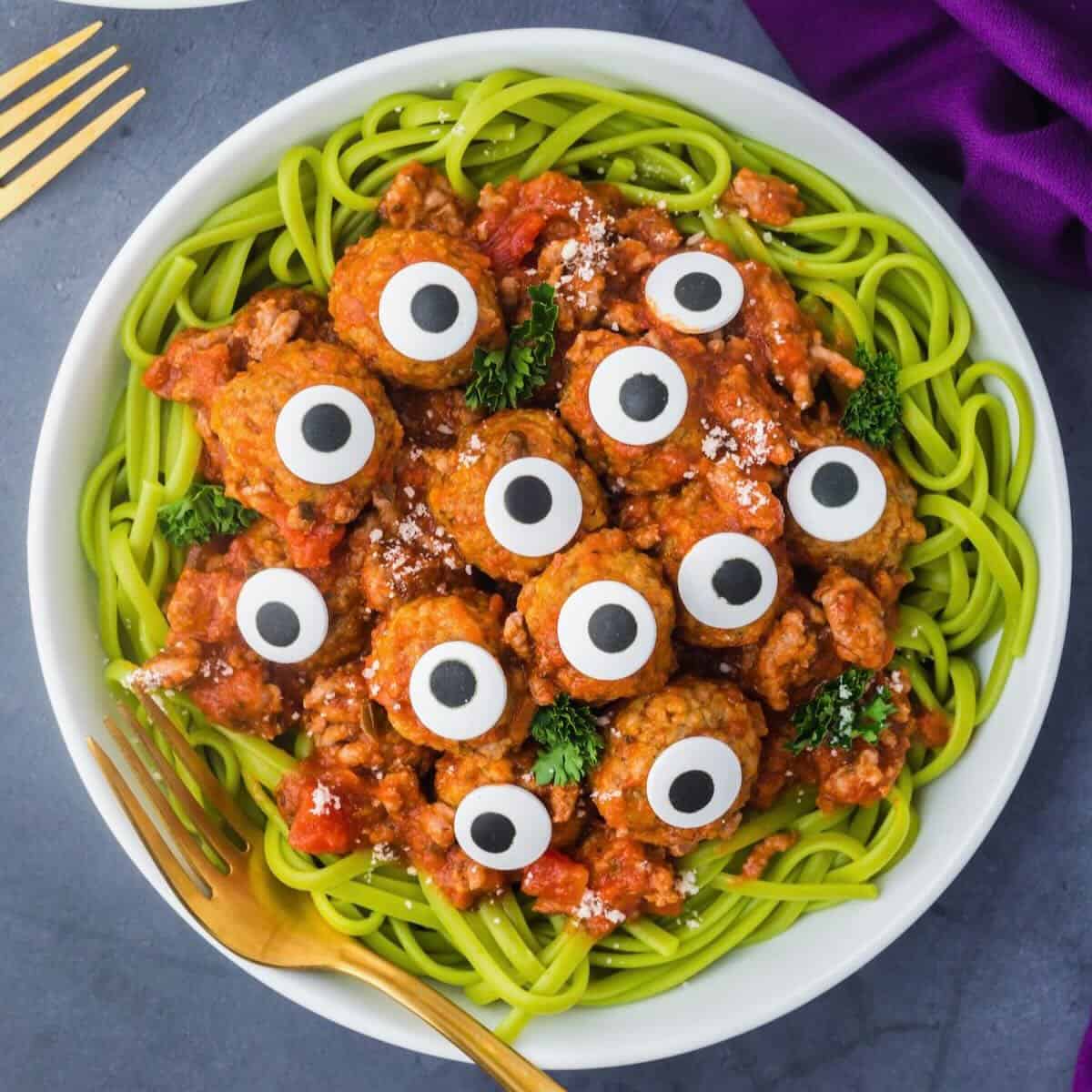 A bowl full of Halloween pasta featuring spinach spaghetti, topped with spaghetti sauce, meatballs, and candy eyes.