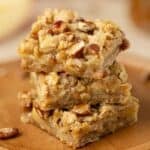 A stack of three oatmeal apple bars with caramel and pecans on a cutting board.