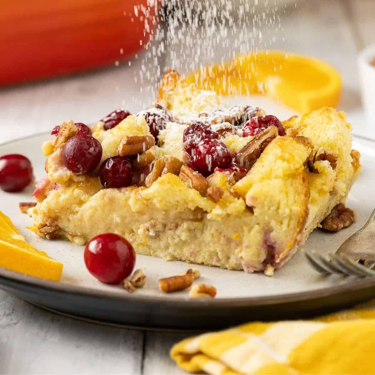A serving of Brioche French Toast Casserole on a plate with powdered sugar being sprinkled over the top.