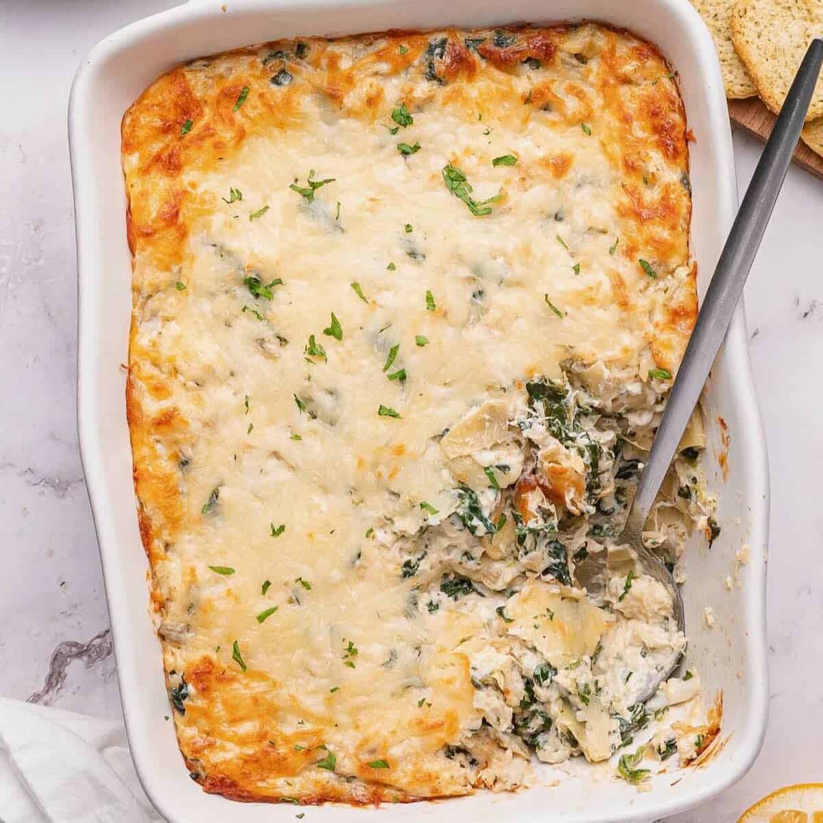 Creamy crab, spinach and artichoke dip in a white rectangle baking dish with a spoonful taken out.
