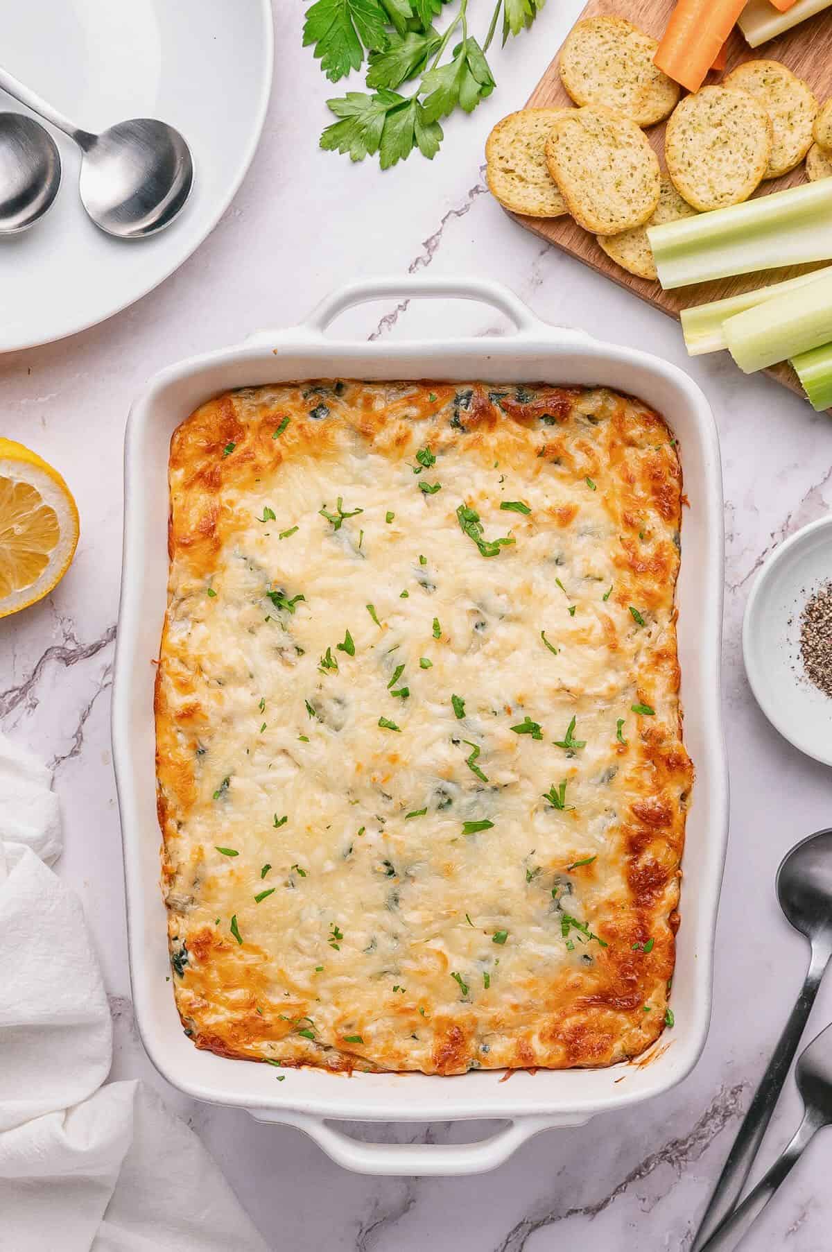 A white baking dish with baked crab, spinach, and artichoke dip garnished with chopped parsley. 
