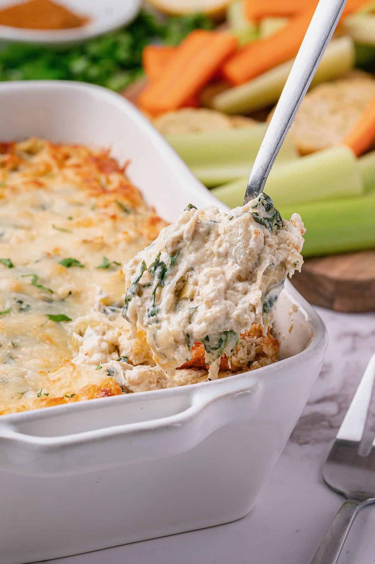A spoonful of baked crab, spinach, and artichoke dip in a white baking dish. 