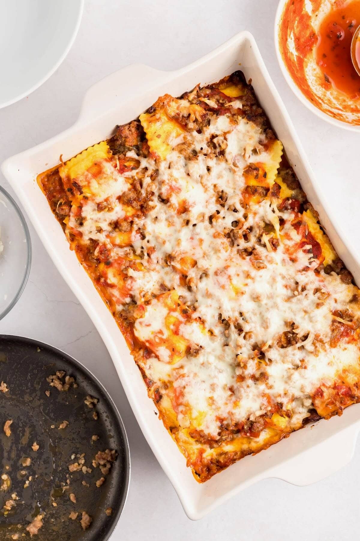 Baked ravioli in a white baking dish with layers of marinara sauce, sausage, ravioli and cheese.
