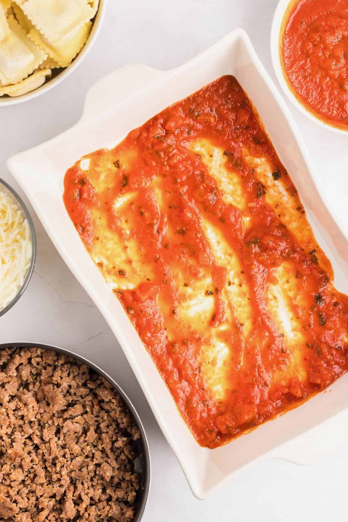 A layer of marinara sauce is spread over the bottom of a white baking dish.