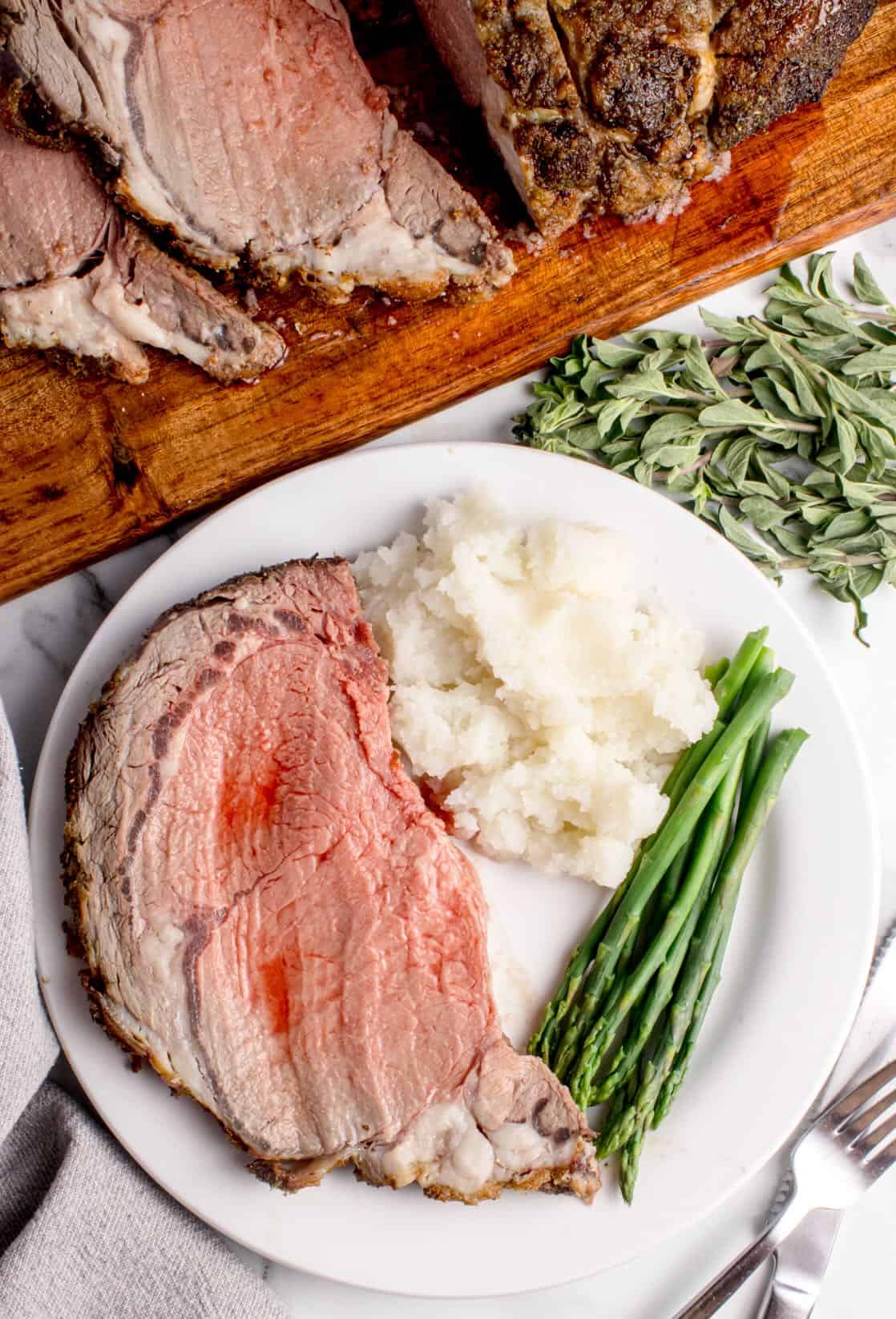 A slice of rare boneless prime rib roast on a plate with mashed potatoes and asparagus. 