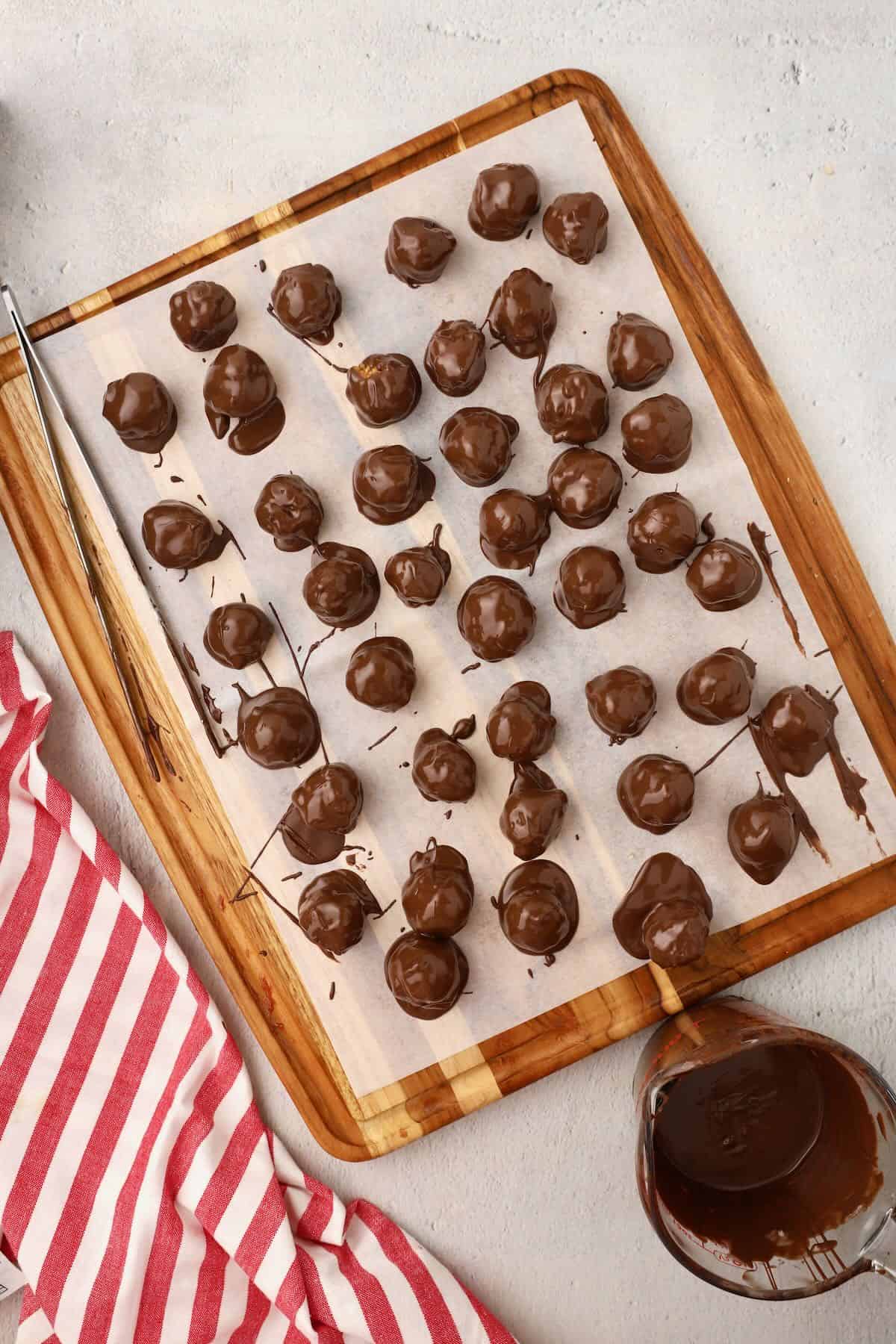 Balls of caramel, rice cereal, and pecans are dipped in melted chocolate and set on parchment paper to for the chocolate to set. 