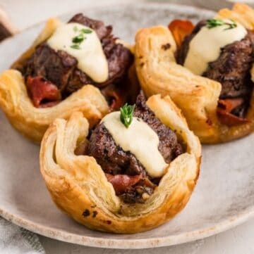 A plate with three mini Beef Wellington Bites topped with horseradish sauce and fresh thyme.