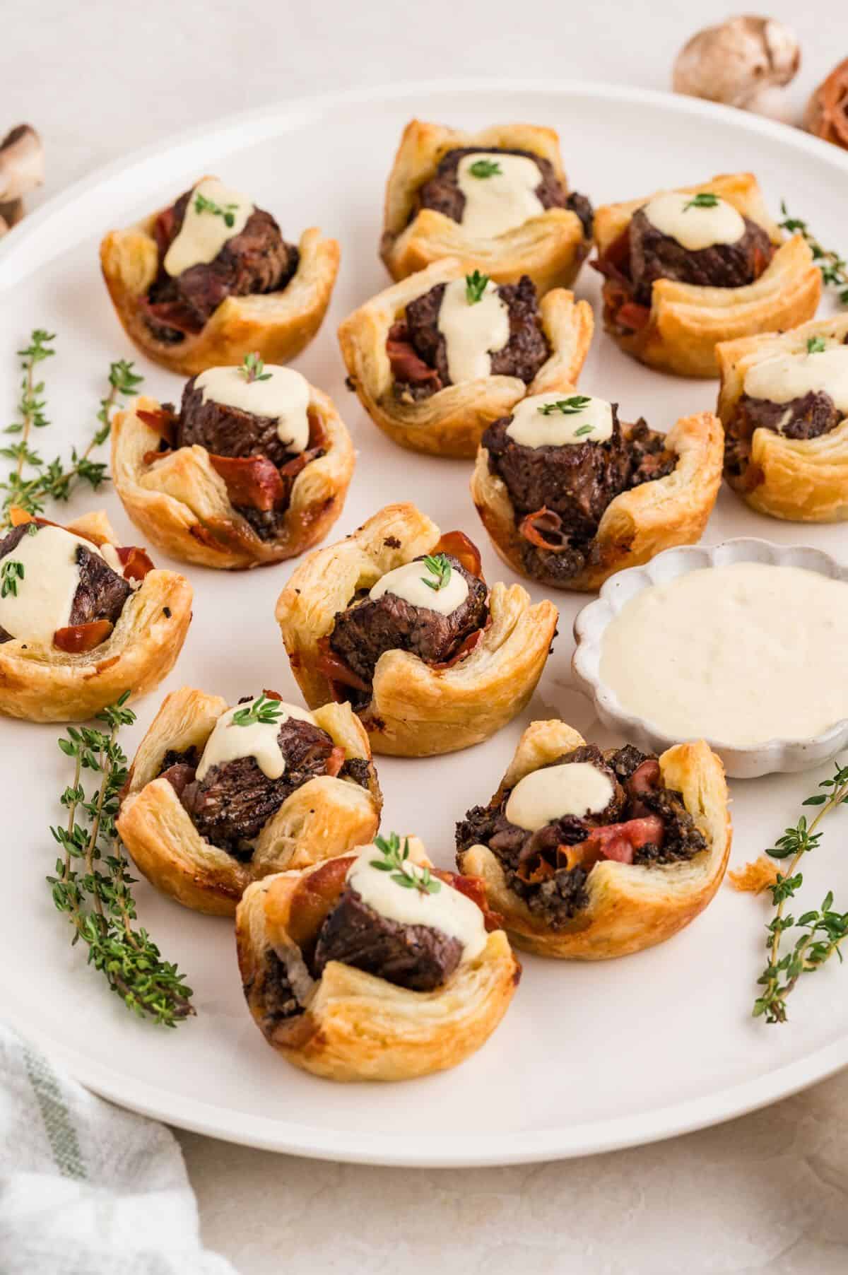 A dozen baked Beef Wellington Bites on a platter garnished with a dollop of horseradish sauce and thyme.