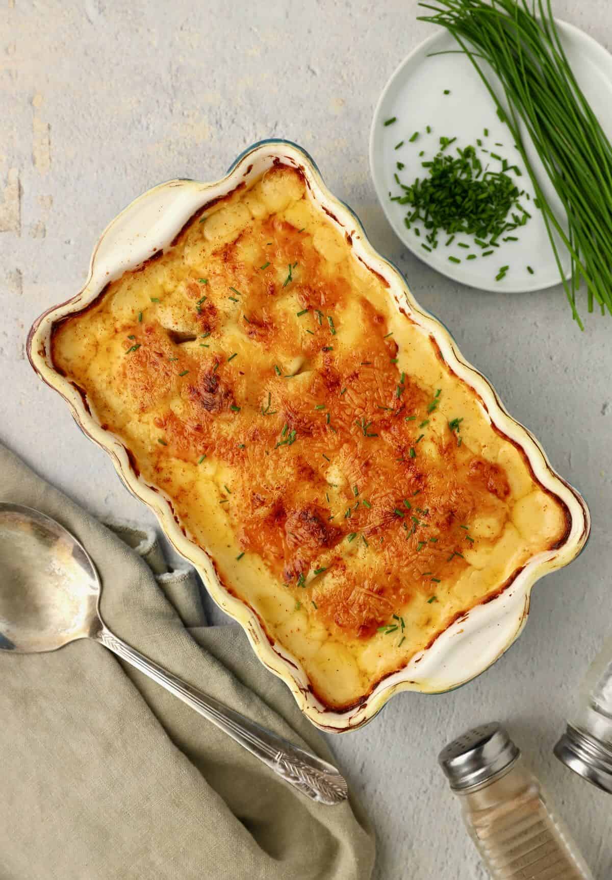 Baked cheesy scalloped potatoes with a plate with chives and a serving spoon.
