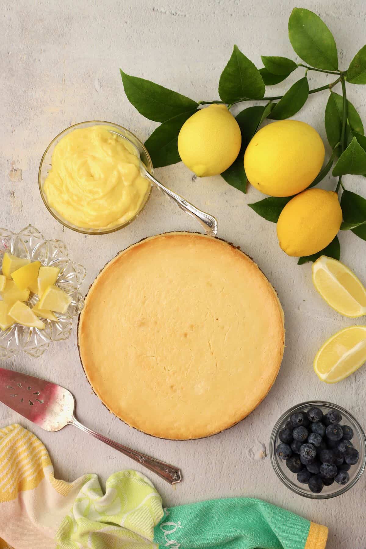 A baked cheesecake with a bowl of lemon curd and fresh lemons.