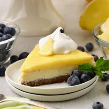 A slice of lemon cheesecake topped with lemon curd on a plate with whipped cream and blueberries.