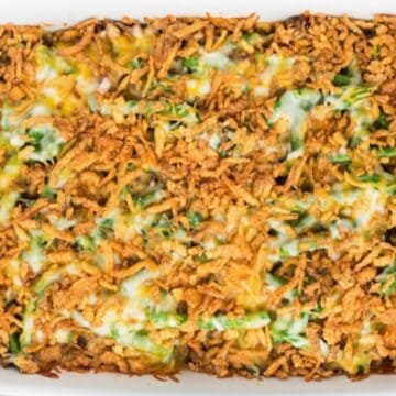 A white casserole dish full of hamburger green bean casserole topped with melted cheese and fried onions.