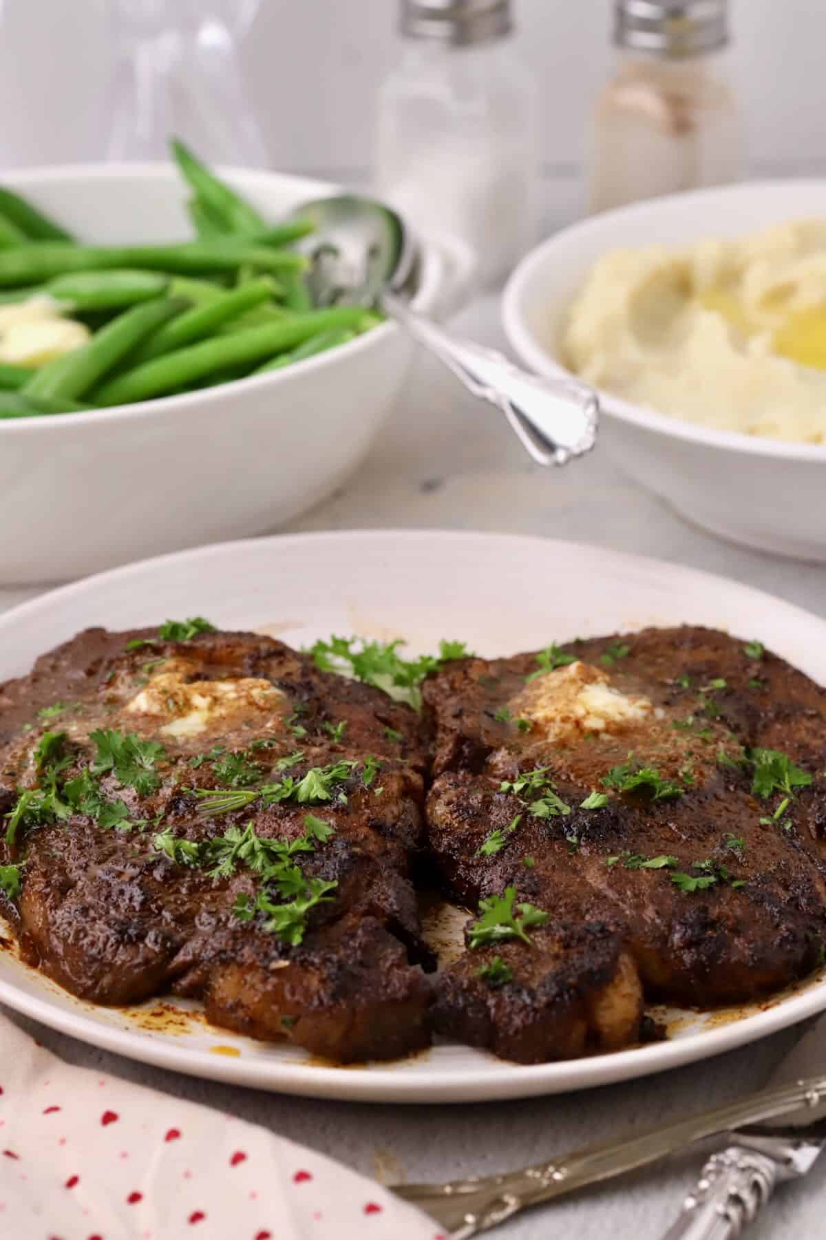 Two pan-seared boneless ribeye steaks topped with pats of maple bourbon butter and chopped parsley on a white plate.