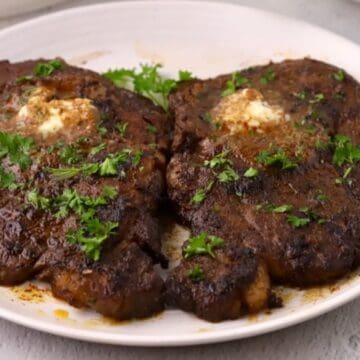 Two pan seared ribeye steaks on a plate topped with bourbon butter and chopped parsley.