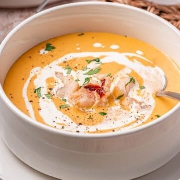 A white bowl full of lobster bisque garnished with pieces of lobster, herbs, and a swirl of cream.