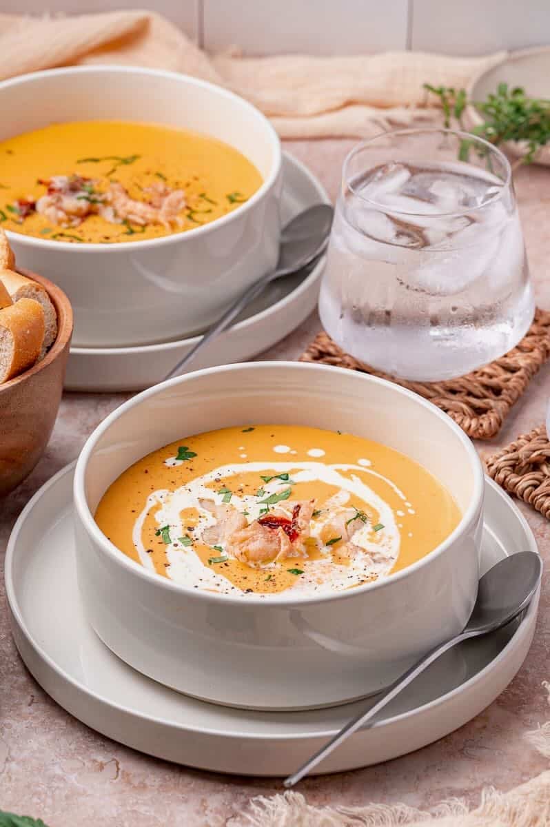 Two bowls of lobster bisque, one garnished with lobster, herbs, and cream.