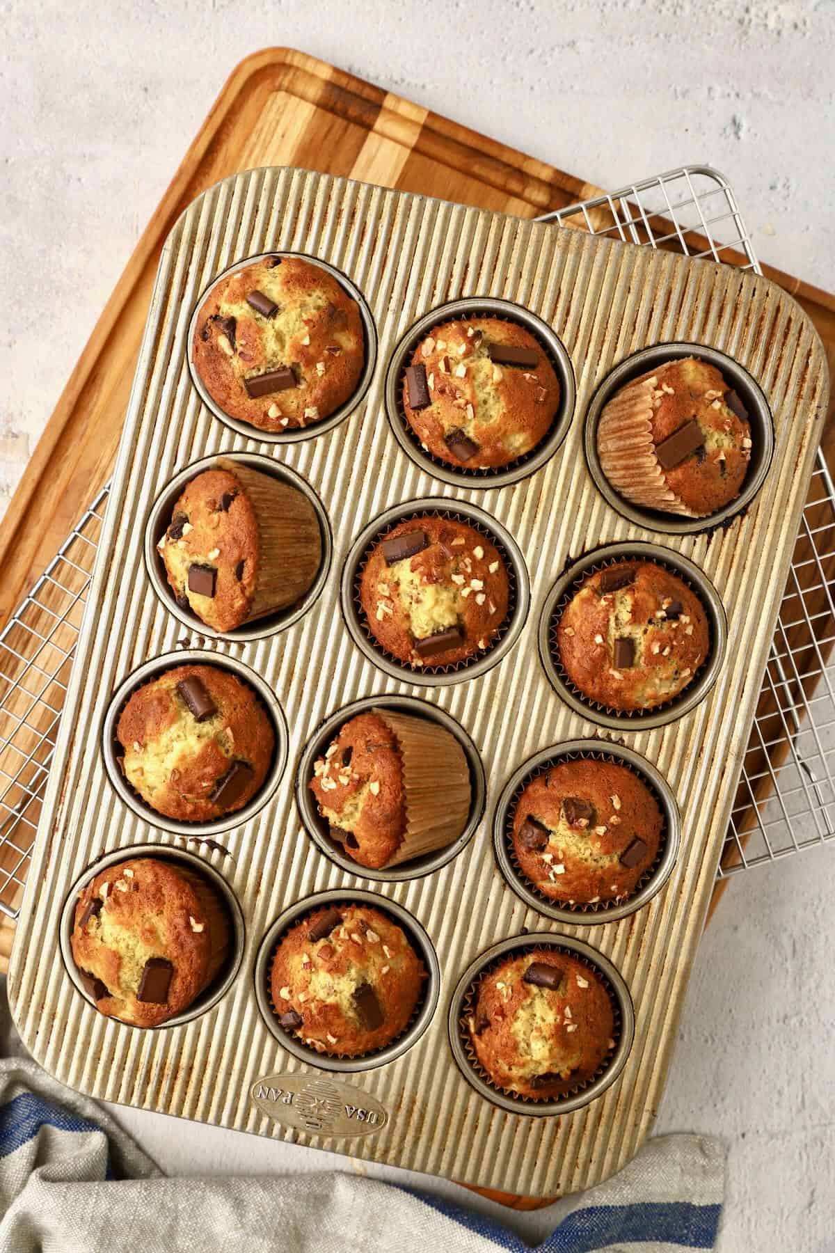 Baked chocolate chunk banana muffins in a muffin tin, cooling on a wire rack.