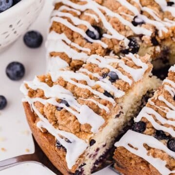 A Lemon Blueberry Coffee Cake topped with blueberries, streusel, and lemon glaze, with a slice cut out.