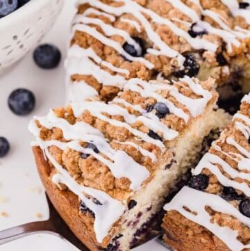 A Lemon Blueberry Coffee Cake topped with blueberries, streusel, and lemon glaze, with a slice cut out.